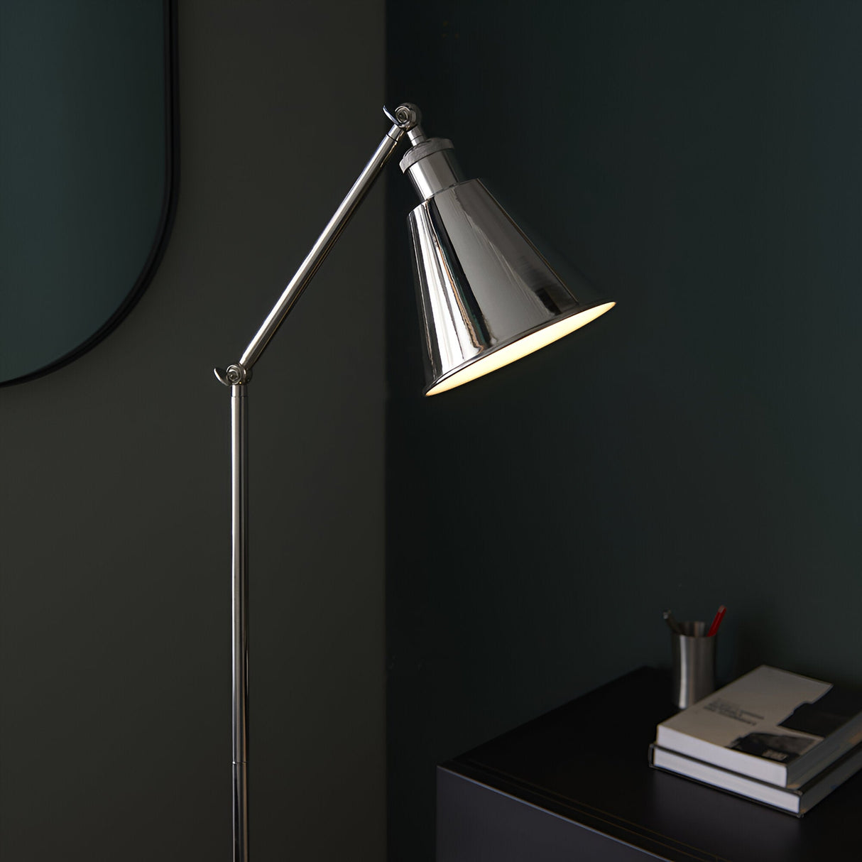 Endon Brackley floor lamp in bright nickel with adjustable arm and conical shade, lit beside a dark green wall with a mirror edge and desk with books.