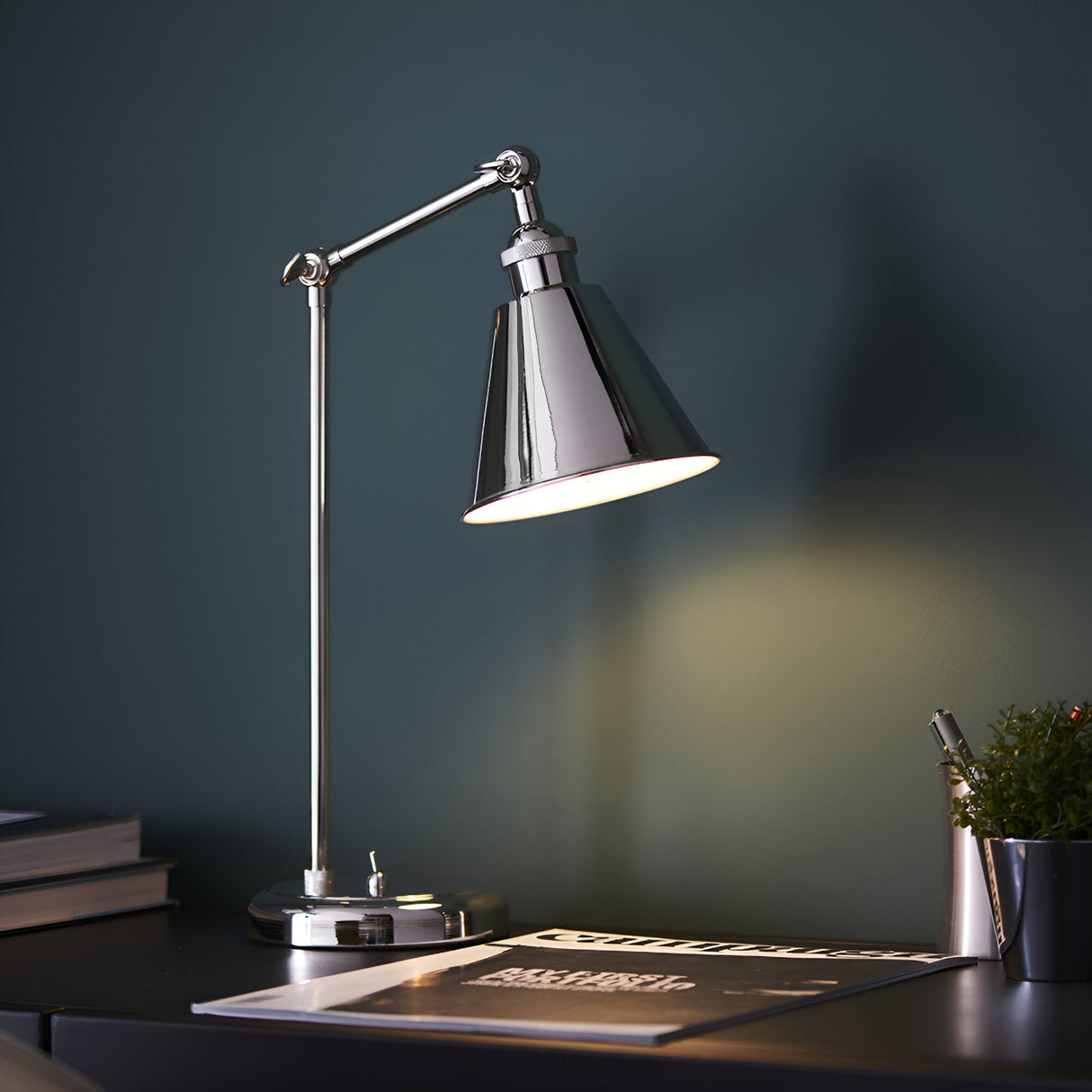 Endon Brackley nickel table lamp with adjustable arm and conical shade illuminated on a desk with books, magazine and plant against a dark teal wall.