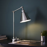 Endon Brackley nickel table lamp with adjustable arm and conical shade illuminated on a desk with books, magazine and plant against a dark teal wall.