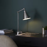 Endon Brackley table lamp in bright nickel with adjustable arm and conical shade, lit on a dark desk with books and a magazine against a teal wall.