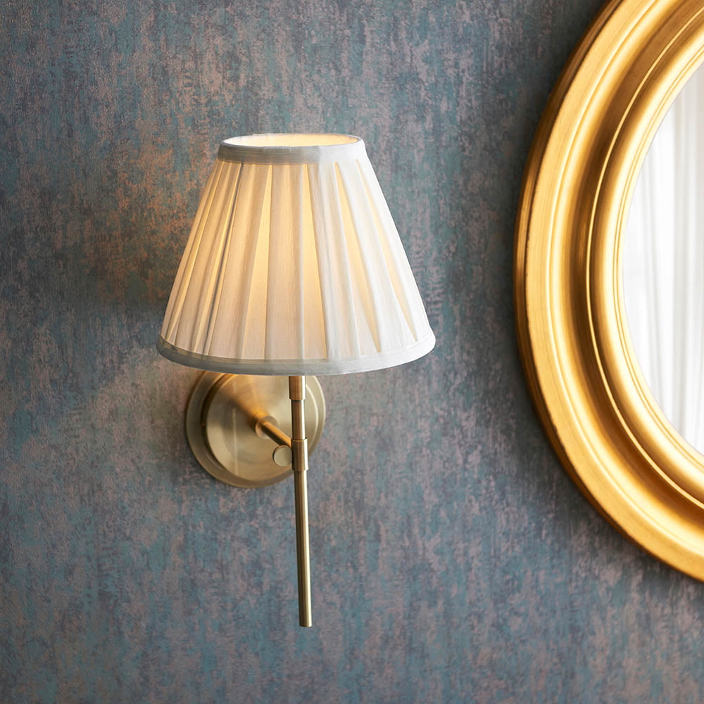 Chartwell Wall Light in brass with a slim metal bracket and pleated cream fabric shade, mounted on a textured blue-grey wall near a round gold-framed mirror.