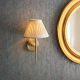 Chartwell Wall Light in brass with a slim upright metal bracket and pleated white fabric shade, mounted beside a round gold-framed mirror.