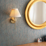 Chartwell Wall Light in brass with pleated fabric shade, mounted on a textured teal wall beside a round gold-framed mirror.