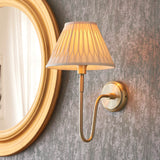 Endon Cliffdon wall light in antique brass with a curved arm and tapered pleated fabric shade, mounted beside a gold oval mirror on a grey wall.