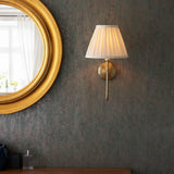 Endon Cliffdon antique brass wall light with a tapered pleated cream shade, mounted on a dark textured wall beside a round gold-framed mirror.