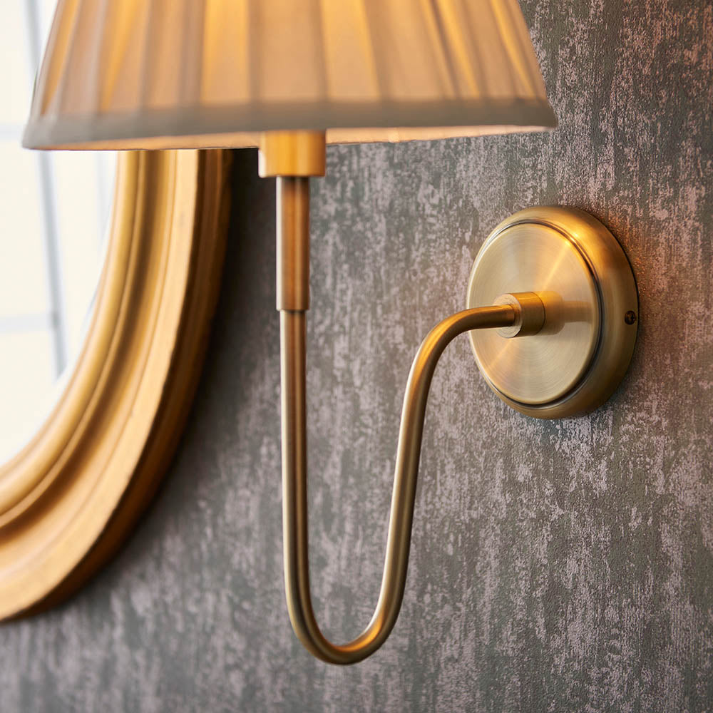 Endon Cliffdon wall light in antique brass, close-up of the curved arm and round backplate mounted beside a gilt mirror on a textured grey wall.