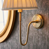 Endon Cliffdon wall light in antique brass, close-up of the curved arm and round backplate mounted beside a gilt mirror on a textured grey wall.