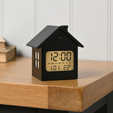 House Digital Alarm Clock with Snooze and Light in black, house-shaped with pitched roof and chimney, LCD display showing time, day and temperature, shown on a wooden desk edge.