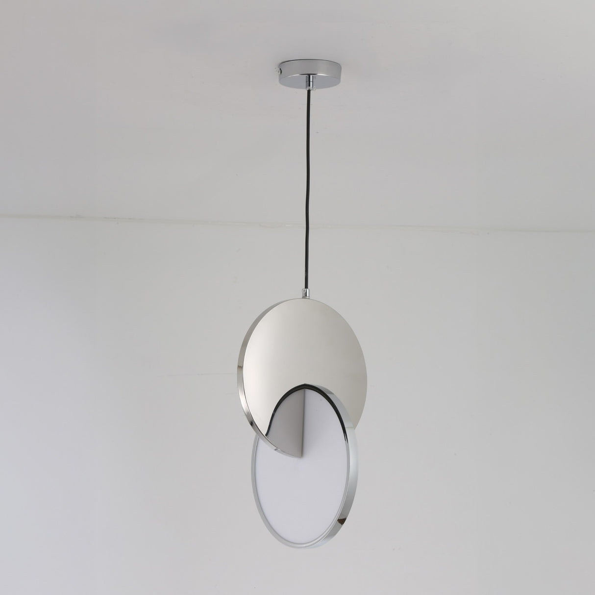 The Half Moon LED Pendant Light - Silver features a circular silver fixture partially covering a flat white diffuser, suspended by a black cord against a plain white ceiling and wall.