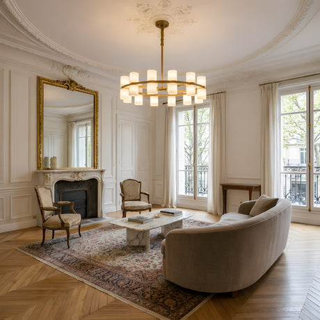 Elegant living room with an ornate ceiling, Valenro 20 Light Cylindrical Alabaster Chandelier in soft white & brass, gold-framed mirror above fireplace, vintage chairs, curved sofa, marble coffee tables, cream curtains, and a patterned rug on parquet floors.