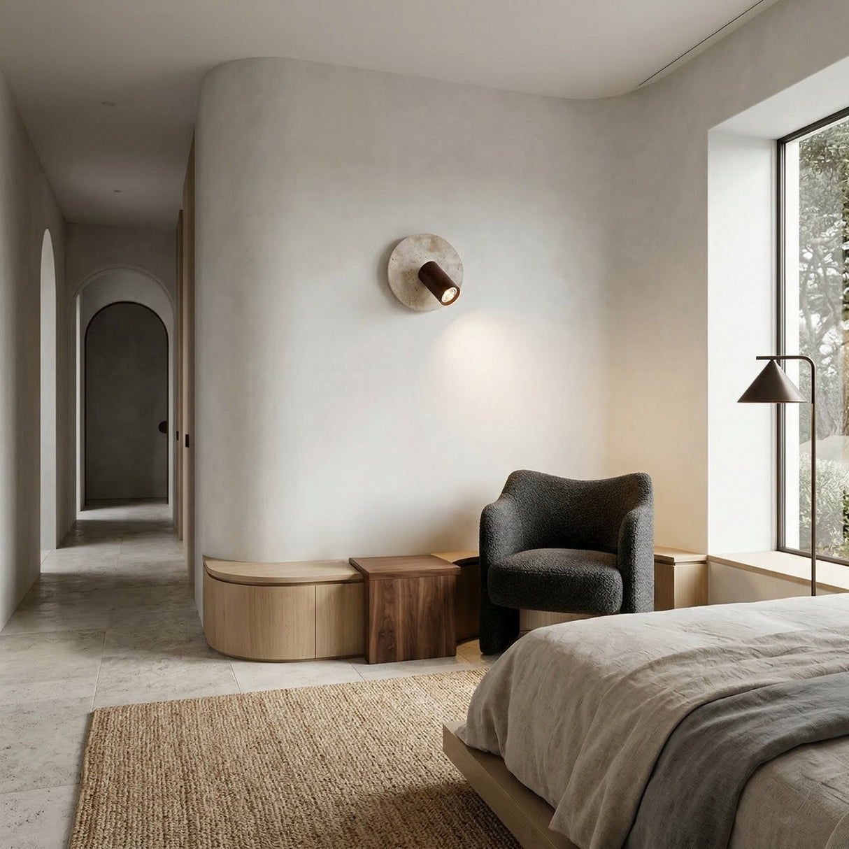 A minimalist bedroom in neutral tones features a large window, cozy dark armchair by a wooden side table, built-in bench, an arched doorway, and textured rug. The Korvane 1 Light Travertine Spotlight in Walnut Brown & Light Beige adds modern warmth.