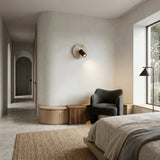 A minimalist bedroom in neutral tones features a large window, cozy dark armchair by a wooden side table, built-in bench, an arched doorway, and textured rug. The Korvane 1 Light Travertine Spotlight in Walnut Brown & Light Beige adds modern warmth.