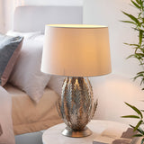 Endon Colette table lamp with silver leaf metal base and ivory cotton drum shade, lit on a bedside table beside a bed and green foliage.