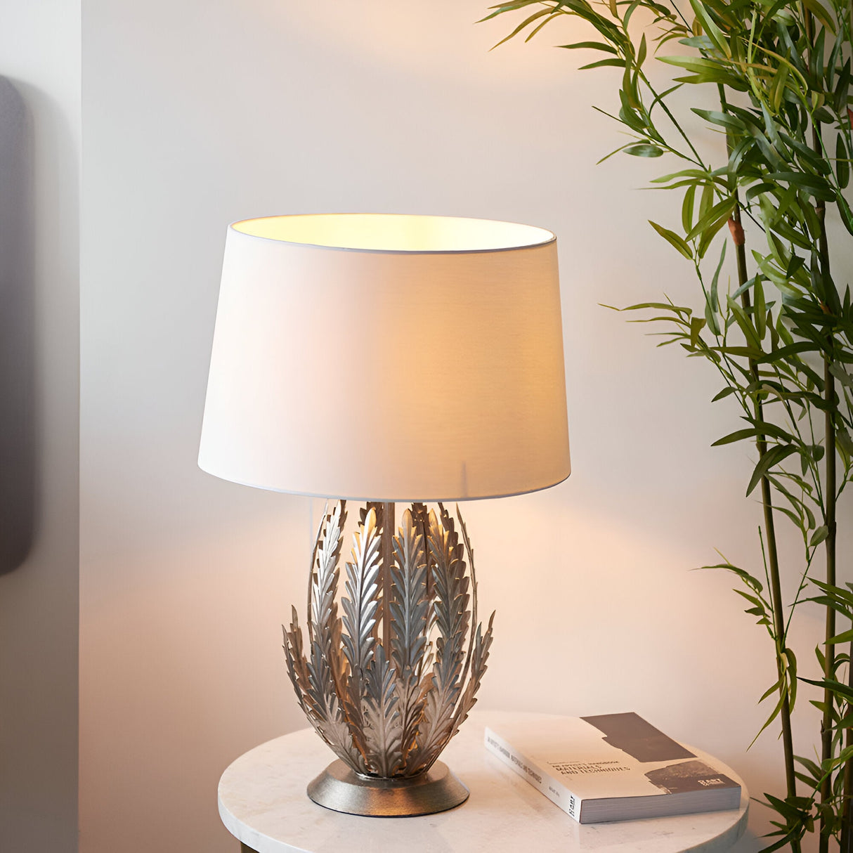 Endon Colette table lamp with layered silver metal leaf base and ivory cotton drum shade, lit on a marble side table beside a bamboo plant.
