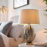Endon Colette table lamp with layered silver leaf metal base and ivory drum shade, lit on a bedside table beside cushions and a framed mirror.
