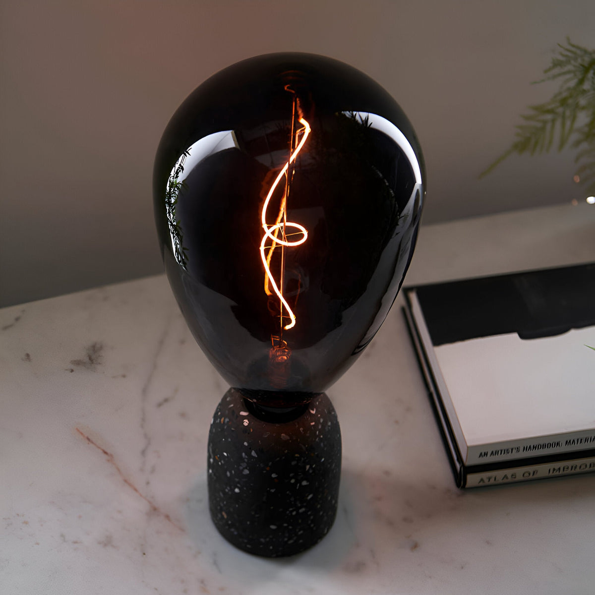 4W E27 LED Filament Wisp Bulb in smoke glass with warm white spiral filament, fitted on a black speckled terrazzo lamp base beside books.