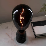 4W E27 LED Filament Wisp Bulb in smoke glass with warm white spiral filament, fitted on a black speckled terrazzo lamp base beside books.