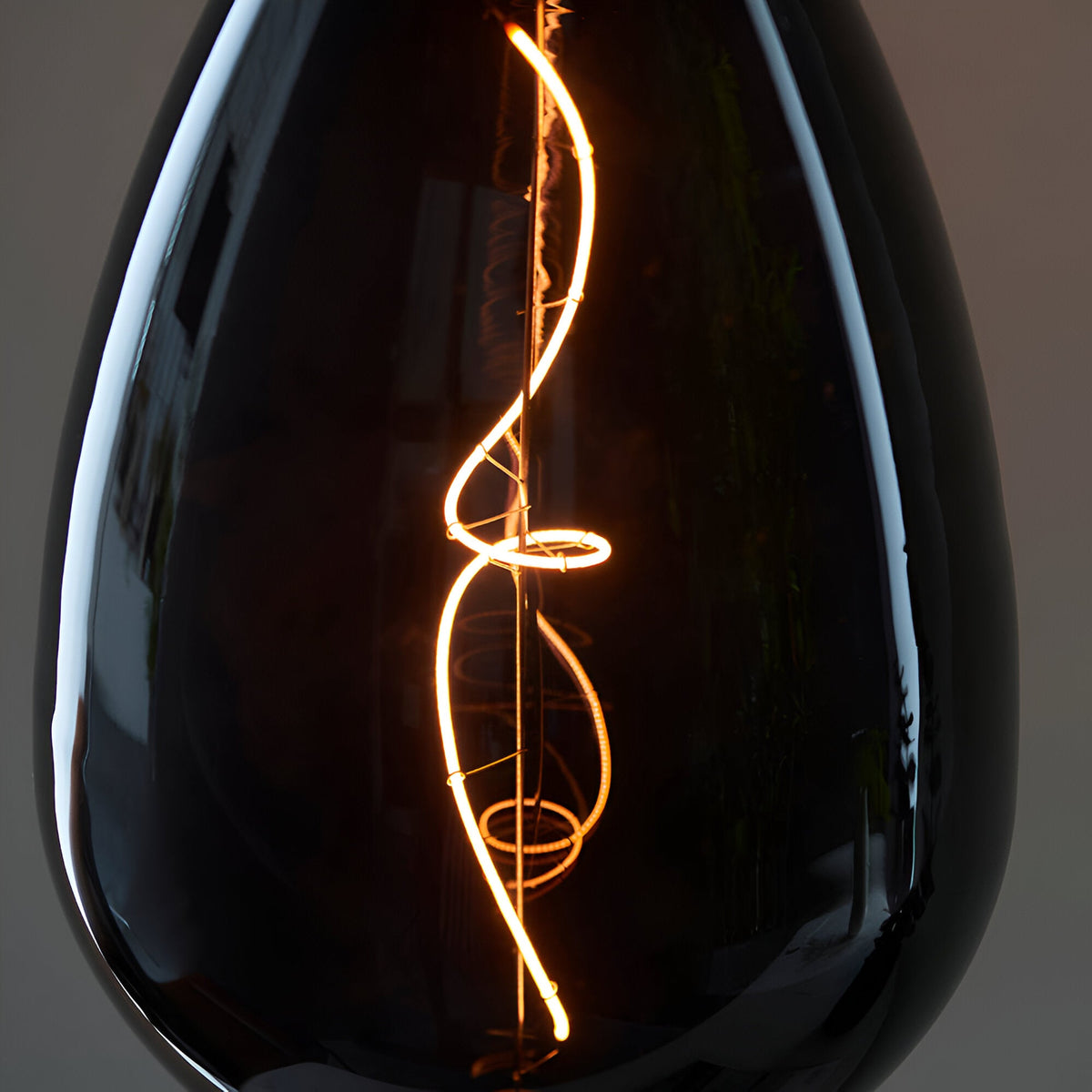 Close-up of the 4W E27 LED Filament Wisp Bulb in smoke glass, showing its spiral warm white filament glowing amber against a dark background.