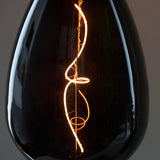 Close-up of the 4W E27 LED Filament Wisp Bulb in smoke glass, showing its spiral warm white filament glowing amber against a dark background.