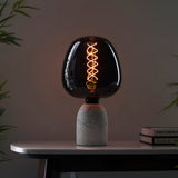 Endon 4W E27 LED filament helix bulb with smoked glass and warm white spiral filament, shown lit in a terrazzo holder on a marble table beside books.