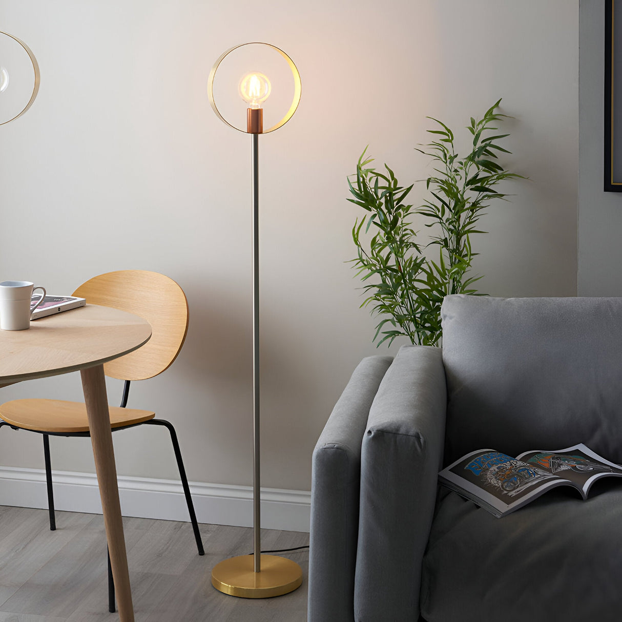 Brenmere Floor Lamp with a round open hoop head, exposed bulb, and brushed brass, copper and nickel finish, shown lit in a living room beside a sofa.