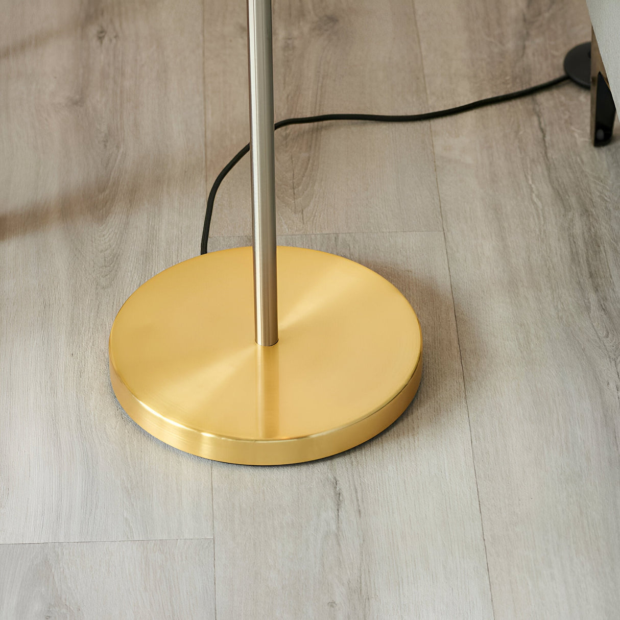Close-up of the Brenmere Floor Lamp's round brushed brass base with nickel-toned pole, black cable trailing across a wood floor.