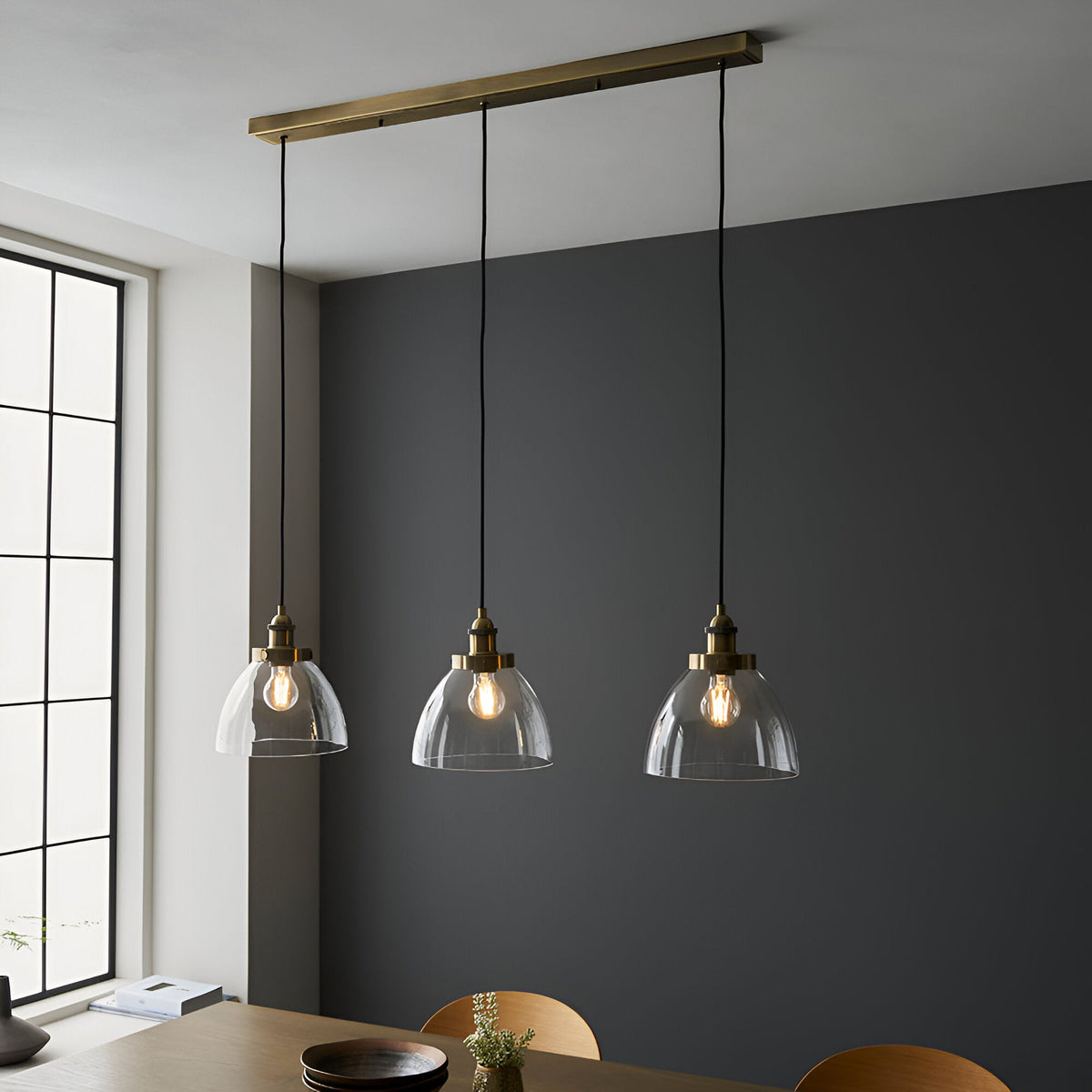 Wentworth 3 Light Linear Pendant in antique brass, three clear glass dome shades on black flex, hung over a dining table by a dark grey wall.