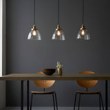 Wentworth 3 Light Linear Pendant in antique brass, clear glass dome shades with visible filament bulbs, hung over a dining table in a dark grey room.