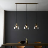 Wentworth 3 Light Linear Pendant in antique brass, with clear glass dome shades on black flex, hung above a dining table against a dark wall.