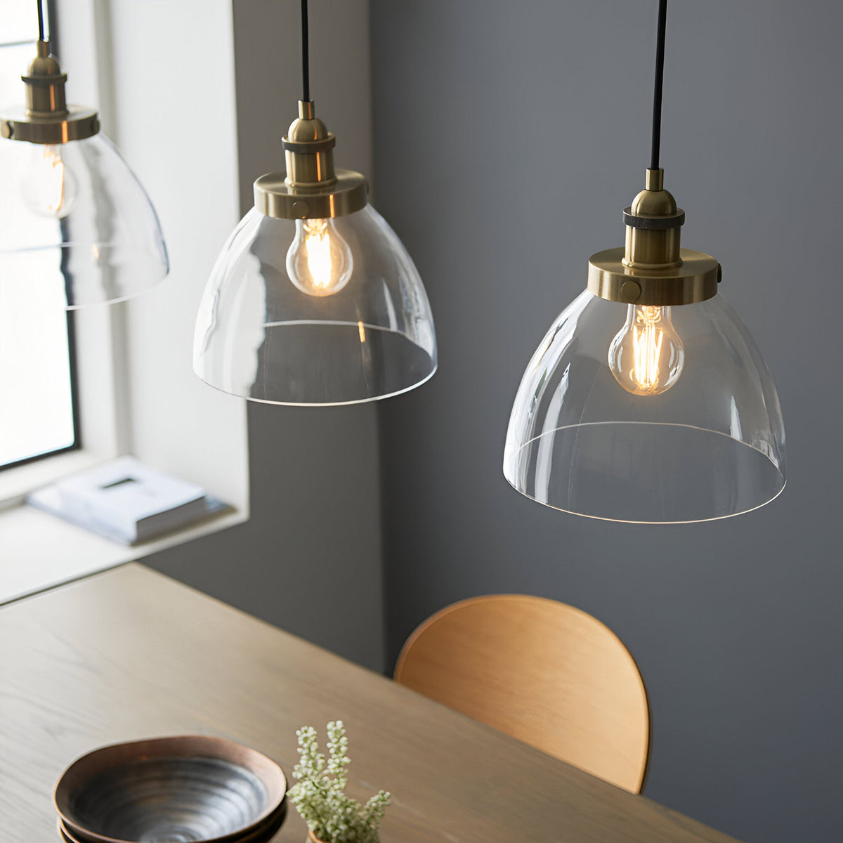 Wentworth 3 Light Linear Pendant in antique brass with clear glass dome shades and exposed filament bulbs, hung over a wooden dining table.
