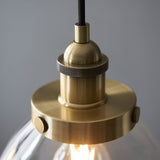 Close-up of the Wentworth 3 Light Linear Pendant in antique brass, showing a knurled E27 lamp holder and clear glass shade against a grey background.