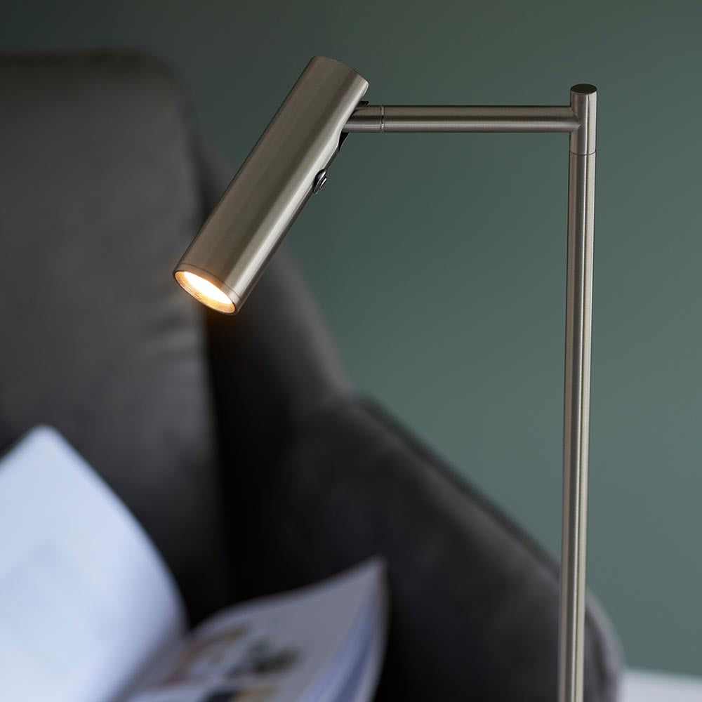 Endon Brackbourne floor lamp in satin nickel with adjustable cylindrical head and push button dimmer, lit beside a dark sofa with an open magazine.
