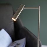 Endon Brackbourne floor lamp in satin nickel with adjustable cylindrical head and push button dimmer, lit beside a dark sofa with an open magazine.
