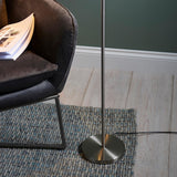 Endon Brackbourne floor lamp base in satin nickel on a woven rug beside a dark velvet chair, with black flex trailing across pale wood flooring.