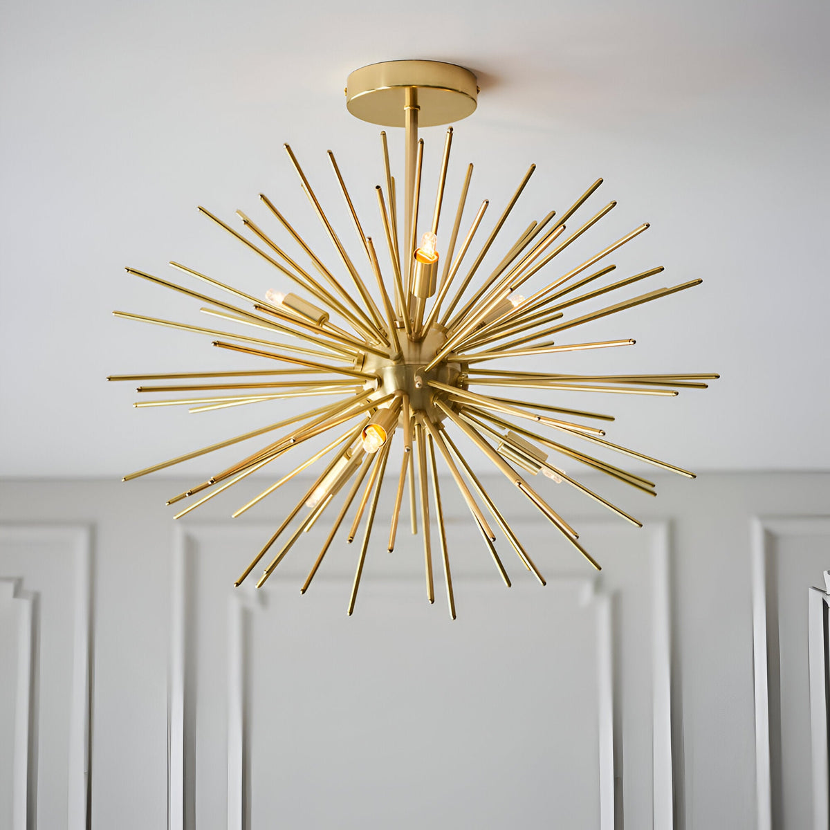 Endon Merrick 6 light semi-flush ceiling light with brass sunburst spike design and exposed G9 lamps, mounted below a white ceiling above panelled walls.