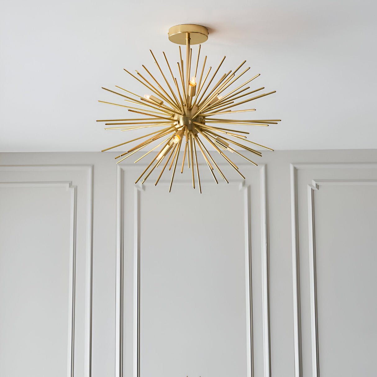Endon Merrick 6 light semi-flush ceiling light in brass, with radiating spike arms forming a starburst, mounted on a panelled white wall interior.