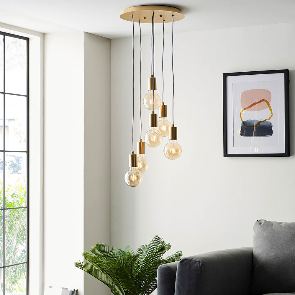 Marlowe 6 Light Pendant in gold, staggered exposed globe bulbs on grey flex cords from a round ceiling plate, shown above a living room seating area.