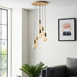 Marlowe 6 Light Pendant in gold, staggered exposed globe bulbs on grey flex cords from a round ceiling plate, shown above a living room seating area.