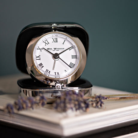 Fold up plastic alarm clock in silver with square case open to reveal a round white Roman numeral dial, shown upright on a book beside dried lavender.