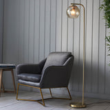 Endon Draycott floor lamp with a champagne dimpled glass globe shade on a brushed brass stand, beside a grey velvet armchair in a living room.