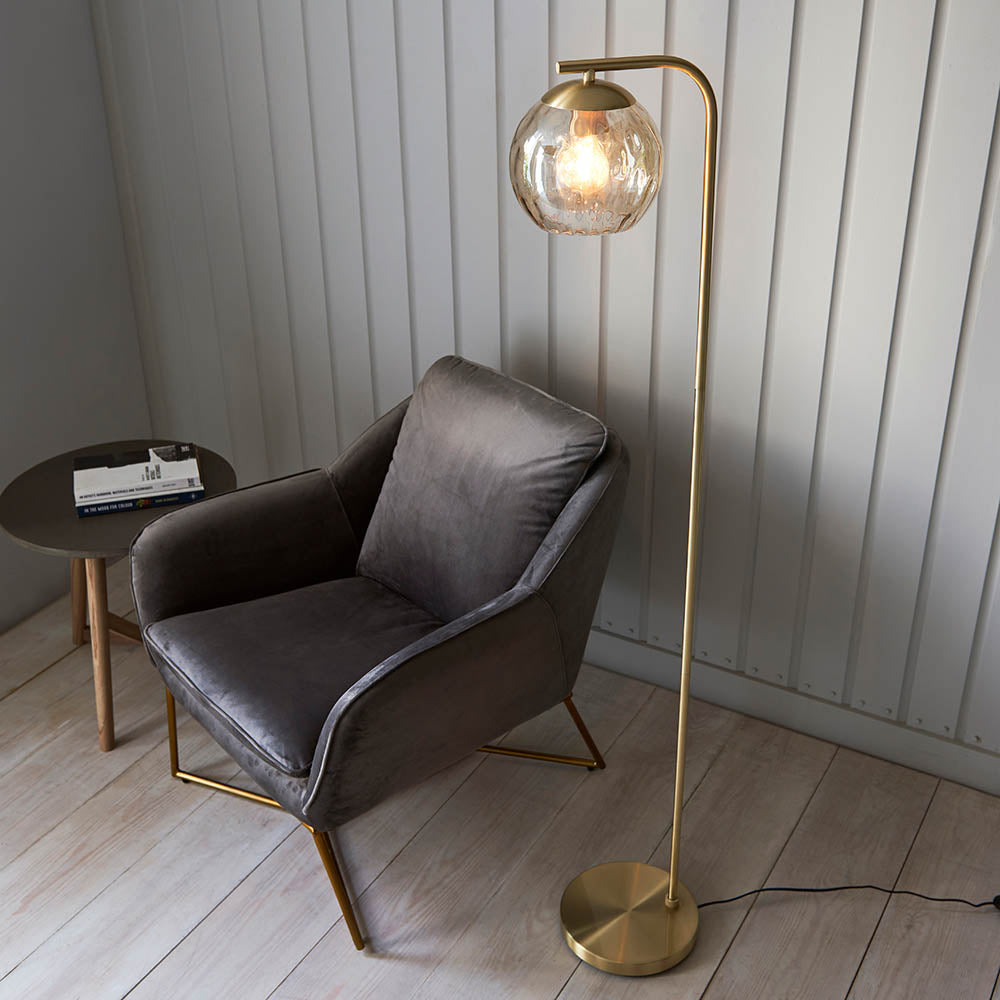 Endon Draycott floor lamp with a dimpled champagne glass globe shade on a brushed brass arched stand, beside a grey velvet armchair and side table.