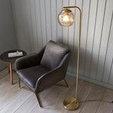 Endon Draycott floor lamp with a dimpled champagne glass globe shade on a brushed brass arched stand, beside a grey velvet armchair and side table.