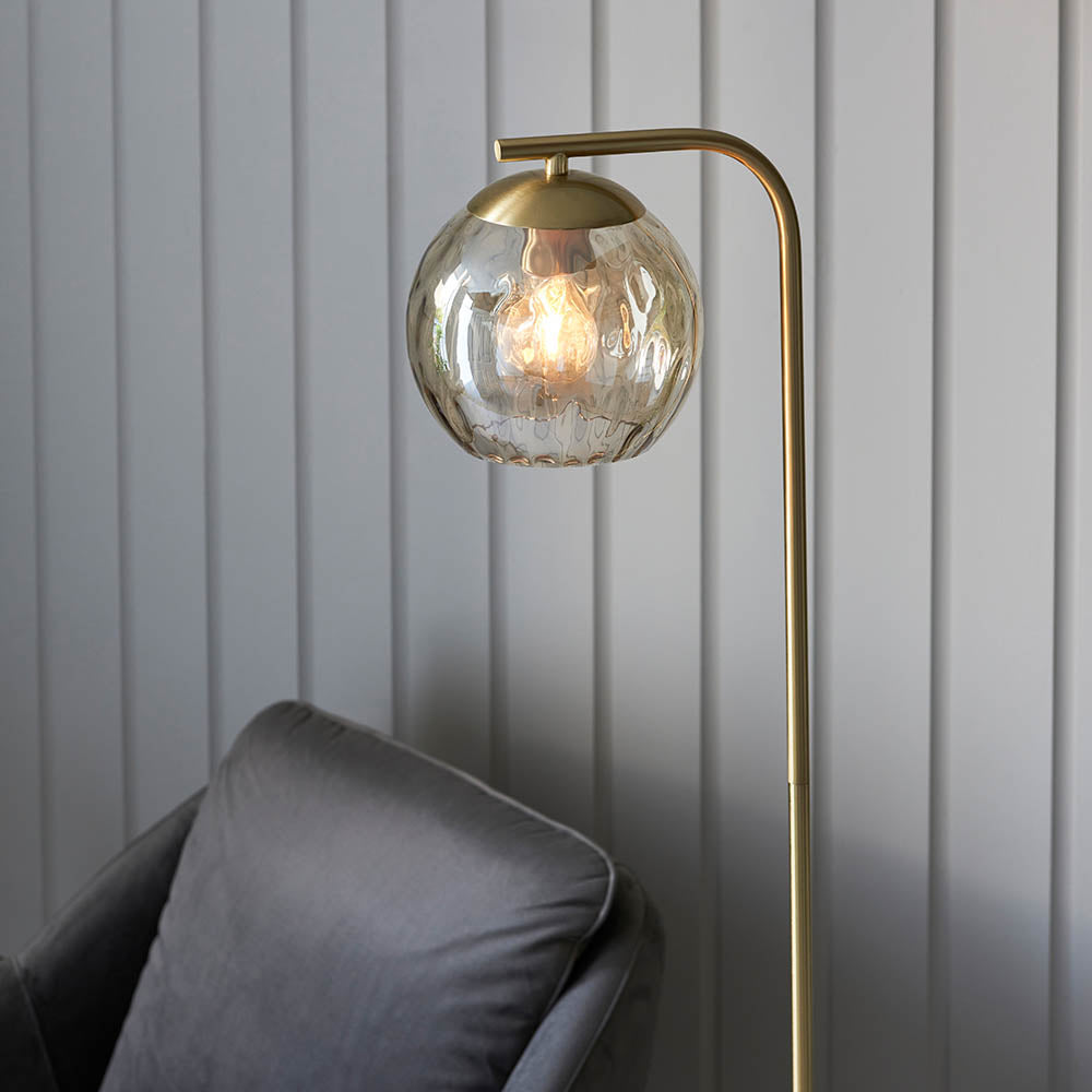 Endon Draycott floor lamp with a champagne dimpled glass globe shade on a brushed brass arched stand, beside a grey velvet sofa and panelled wall.