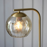 Endon Draycott floor lamp detail with a champagne dimpled glass globe shade and lit E27 filament bulb on a brushed brass arm against grey panelling.