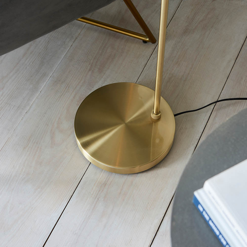 Close-up of the brushed brass weighted round base of the Endon Draycott floor lamp on pale wood planks, with black flex trailing to the side.