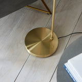Close-up of the brushed brass weighted round base of the Endon Draycott floor lamp on pale wood planks, with black flex trailing to the side.