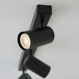 Calder 3 Light Spotlight Plate in matt black metal, close-up of one cylindrical GU10 head lit warm white, angled on a light grey background.