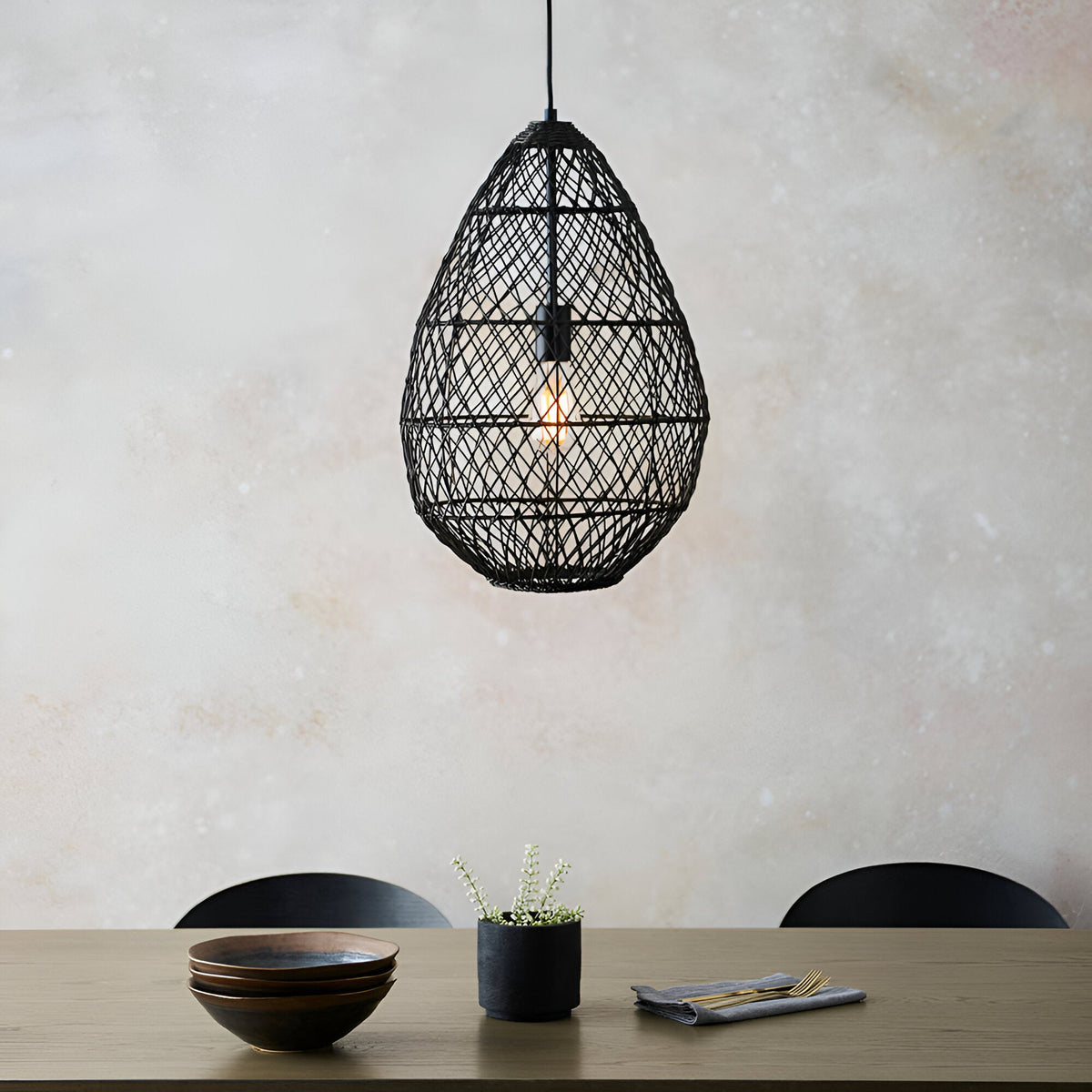 Endon Chartdale teardrop pendant in black woven rattan with a single E27 bulb, hung above a wooden dining table set with bowls and a potted plant.