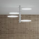 SuperMoon LED semi-flush ceiling light with white vertical stem and four staggered arms holding round crescent-cut discs, mounted on a plain ceiling above a wood-panelled wall.