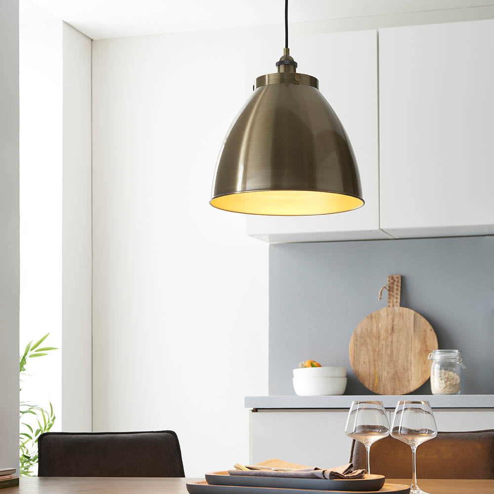 Endon Bramdale medium pendant in antique brass with a domed metal shade on a black flex, hanging above a dining table in a bright modern kitchen.